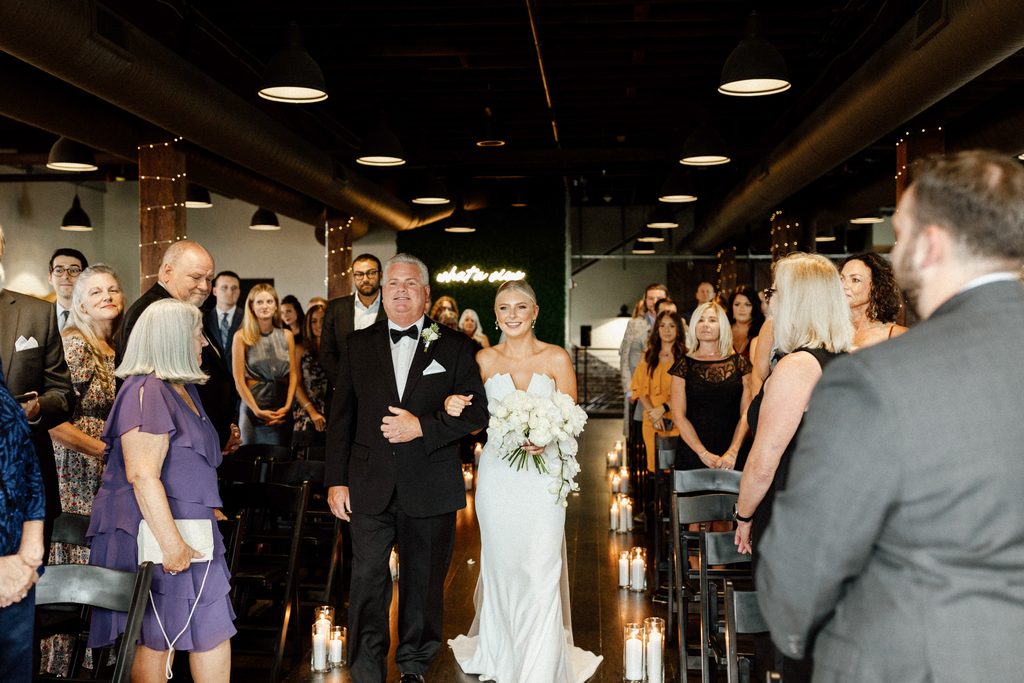 Bride Samantha walks down the aisle with her father during her indoor wedding ceremony at Tinker House Events in Indianapolis, Indiana.