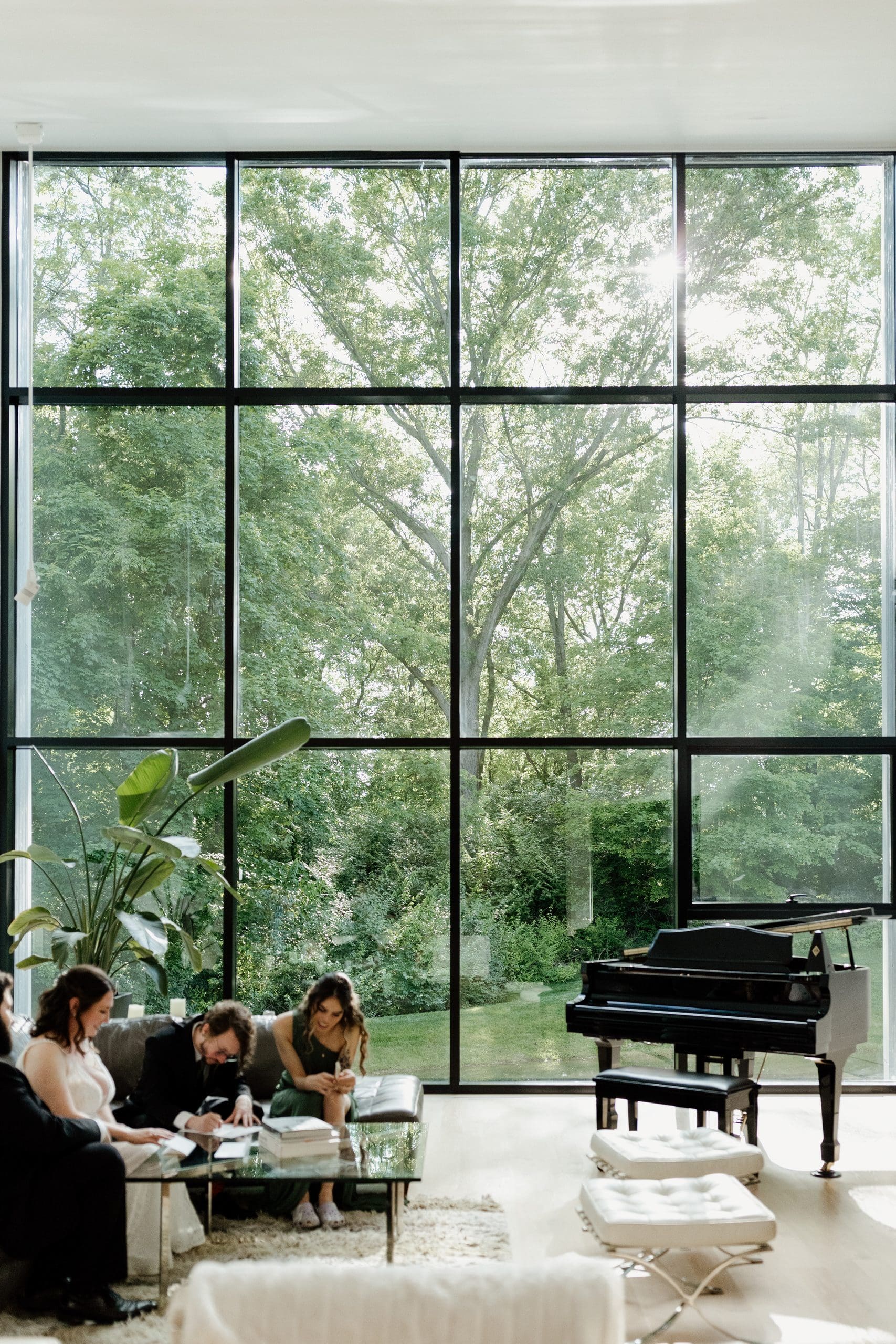 Guests gather inside The Plus House in Indianapolis with floor-to-ceiling windows, garden views and a grand piano during a wedding celebration.