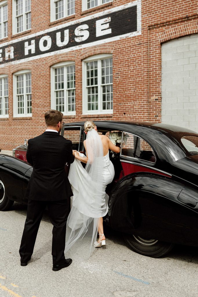Groom Ryan helping bride Samantha into a vintage car during their wedding portraits in historic Tinker House Events, Indianapolis.